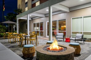 Een terras buiten met tafels en stoelen, een vuurkorf, en een gebouw met grote ramen.