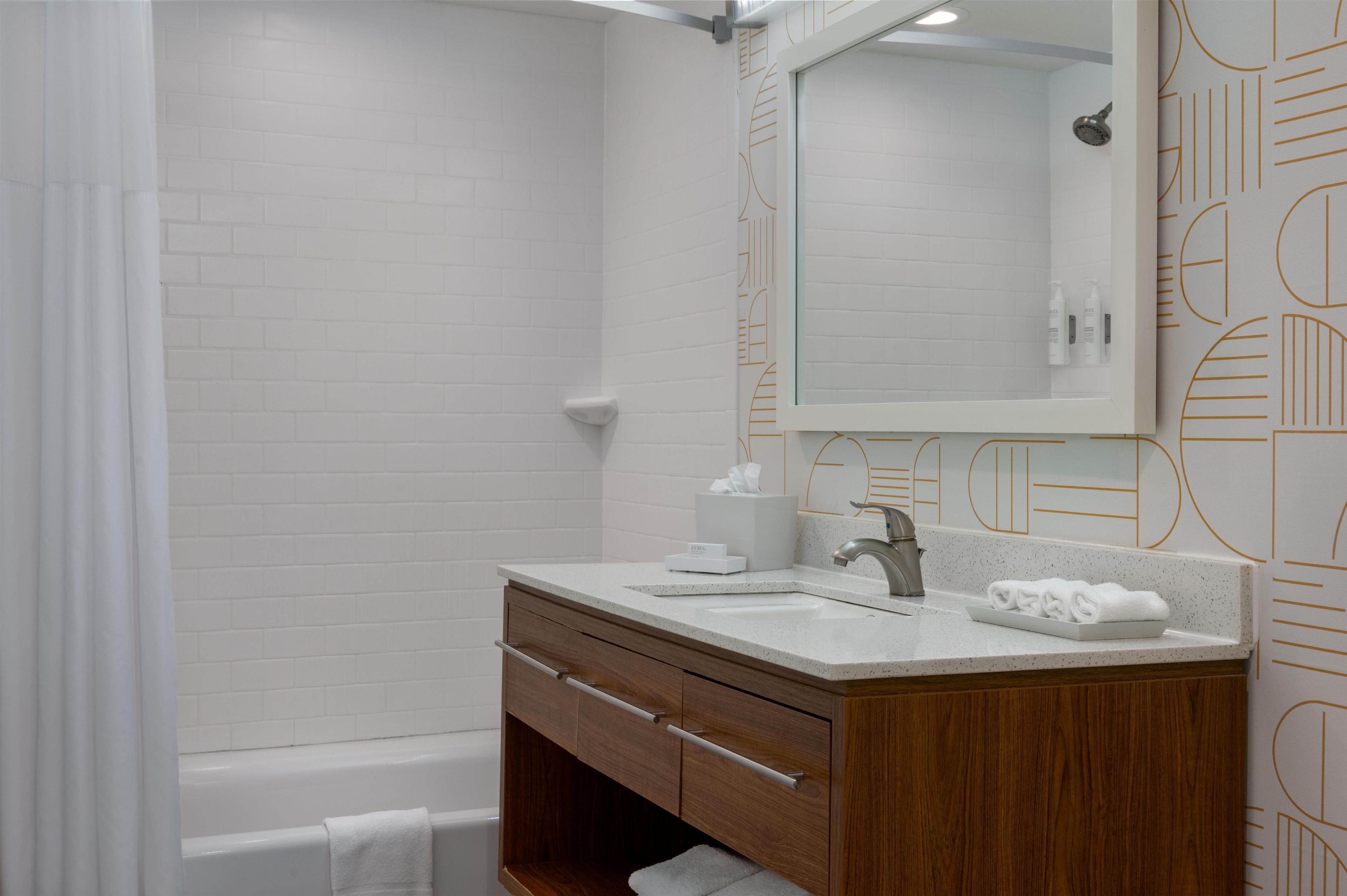 Un baño moderno con ducha de azulejos blancos, tocador de madera con lavamanos y un espejo grande.