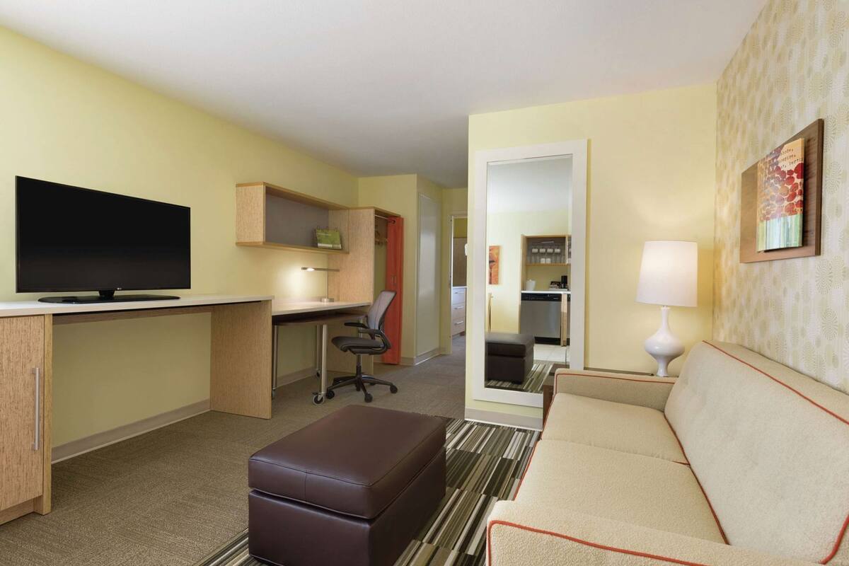 A hotel room with a TV, desk, office chair, sofa, ottoman, and a mirror.