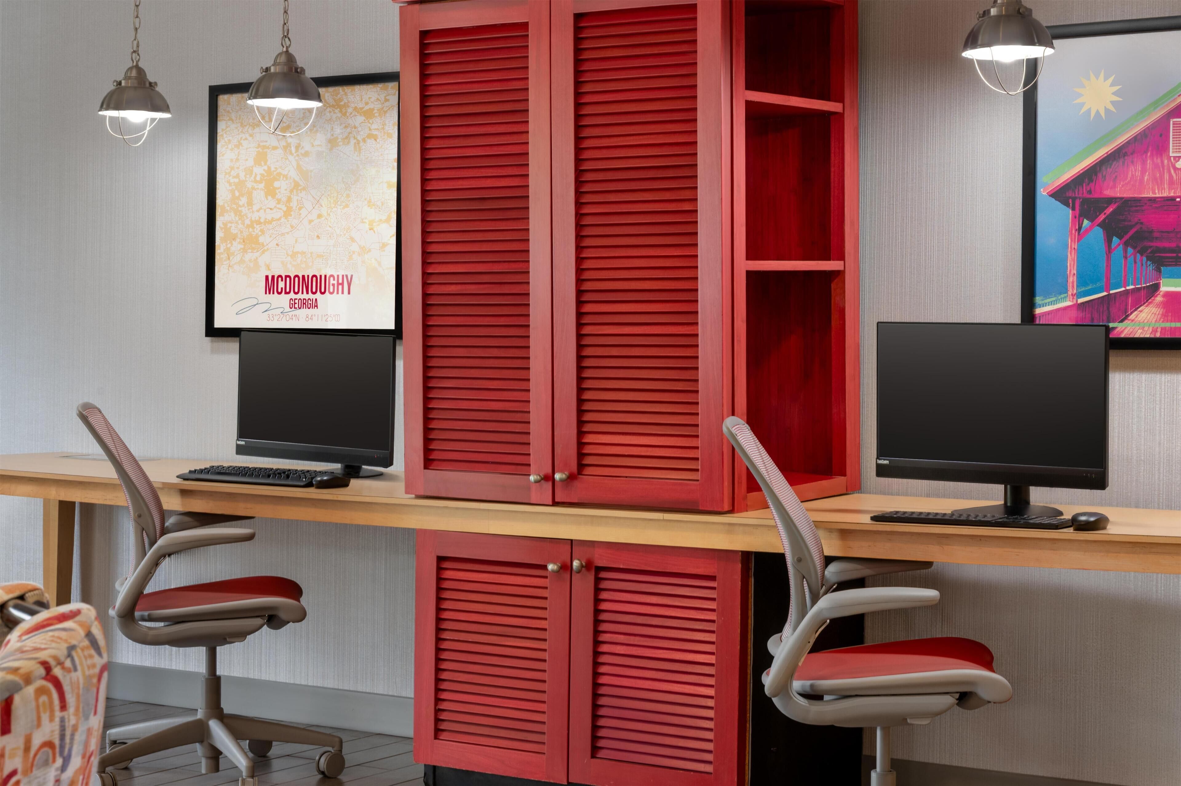 A workspace with two desks, each with a computer monitor, keyboard, and mouse. There are two office chairs with red cushions. The room has a wooden desk and a red cabinet with sliding doors.
