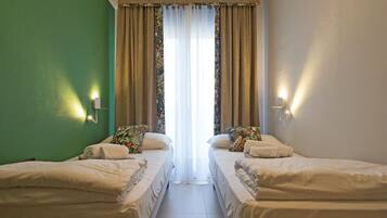 A hotel room with two single beds, a green wall, a large window with curtains, and wall-mounted lights.
