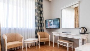 A hotel room with a wooden floor, two beige armchairs, a small white table, a white desk with a computer and a kettle, and a large window with sheer curtains.