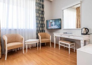 A hotel room with a wooden floor, two beige armchairs, a small white table, a white desk with a computer and a kettle, and a large window with sheer curtains. - Armas Pemar Beach (Manavgat)