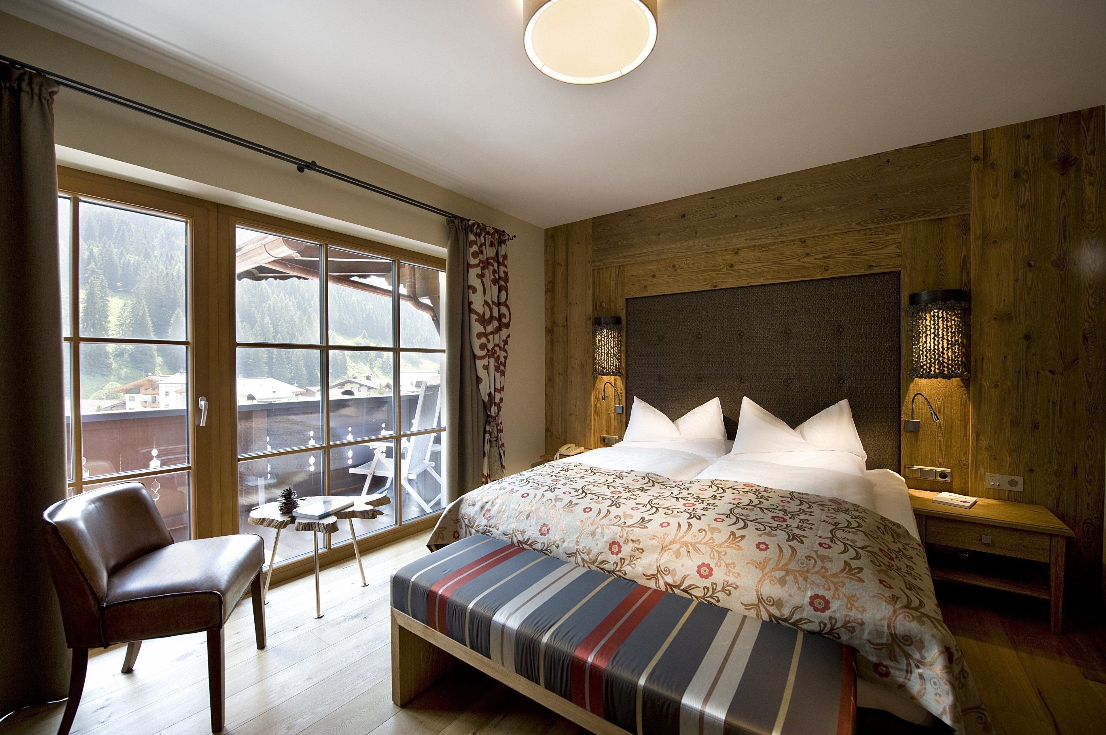 A bedroom with a large window, a bed with a patterned duvet, a bench, a chair, and a small table.