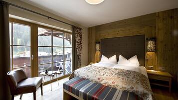 A bedroom with a large window, a bed with a patterned duvet, a bench, a chair, and a small table.