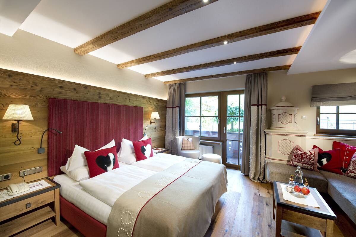 A hotel room with a wooden ceiling, a large bed with red and white bedding, a sitting area with a table and chairs, and a view of the outdoors through a window.