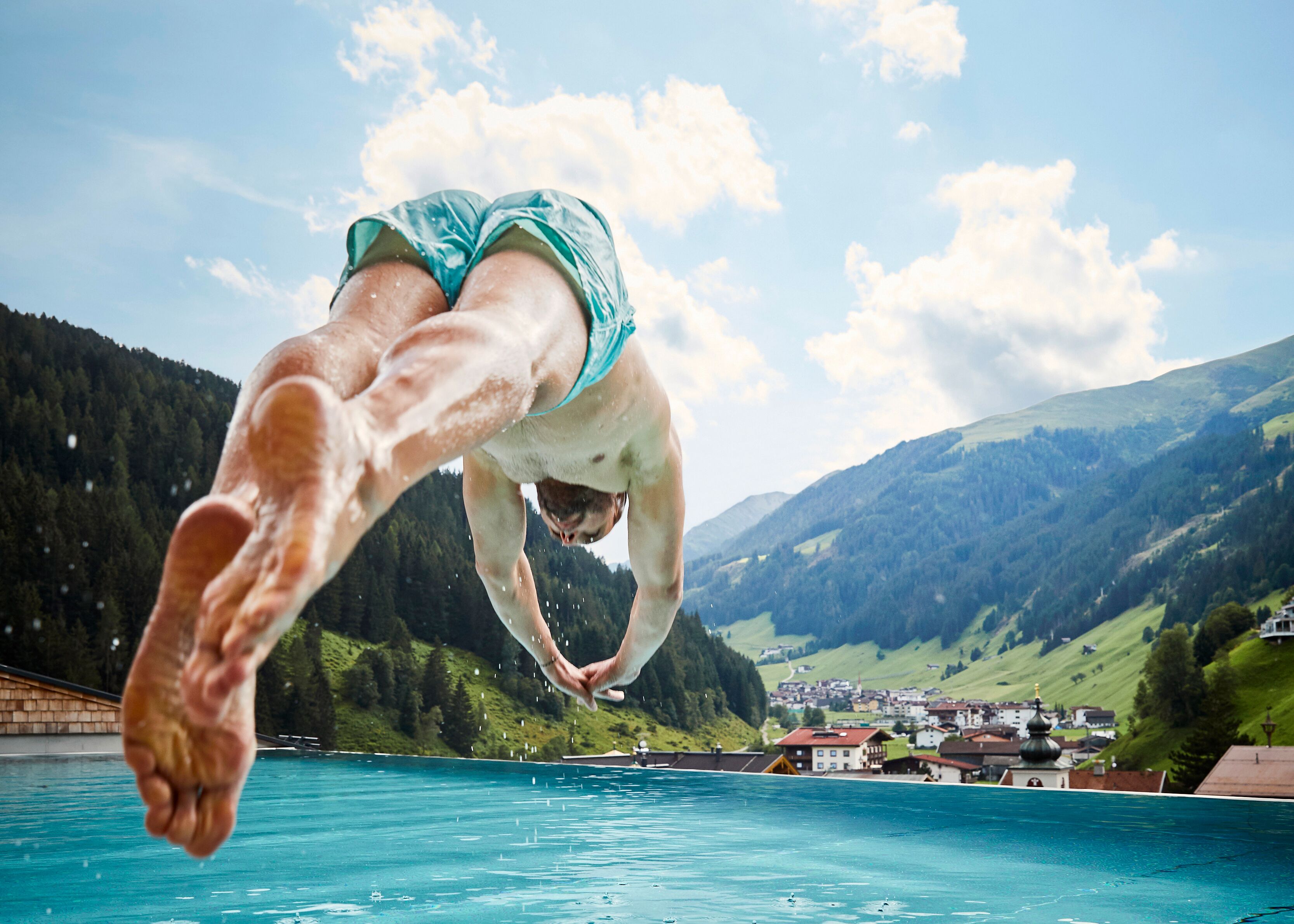 Ein Taucher in der Luft über einem klaren, blauen Pool, im Hintergrund eine gebirgige Landschaft.