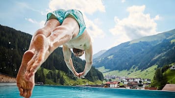 Ein Taucher in der Luft über einem klaren, blauen Pool, im Hintergrund eine gebirgige Landschaft.