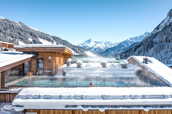 Una piscina termal con una terraza de madera, rodeada de montañas nevadas y un cielo despejado.