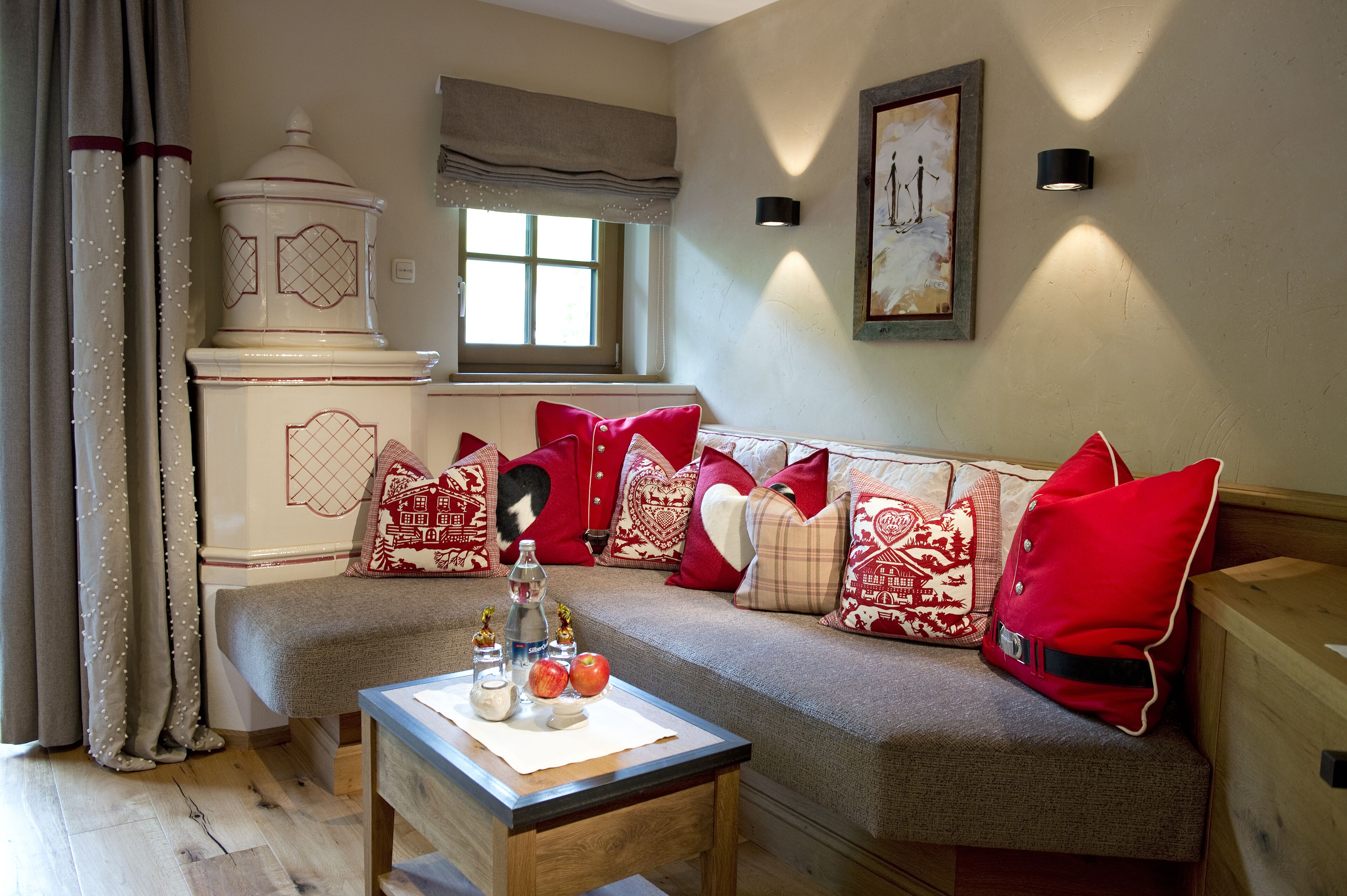 A cozy living room with a sofa adorned with red and white patterned cushions, a wooden coffee table with apples and a bottle, and a window with a view of the outdoors.
