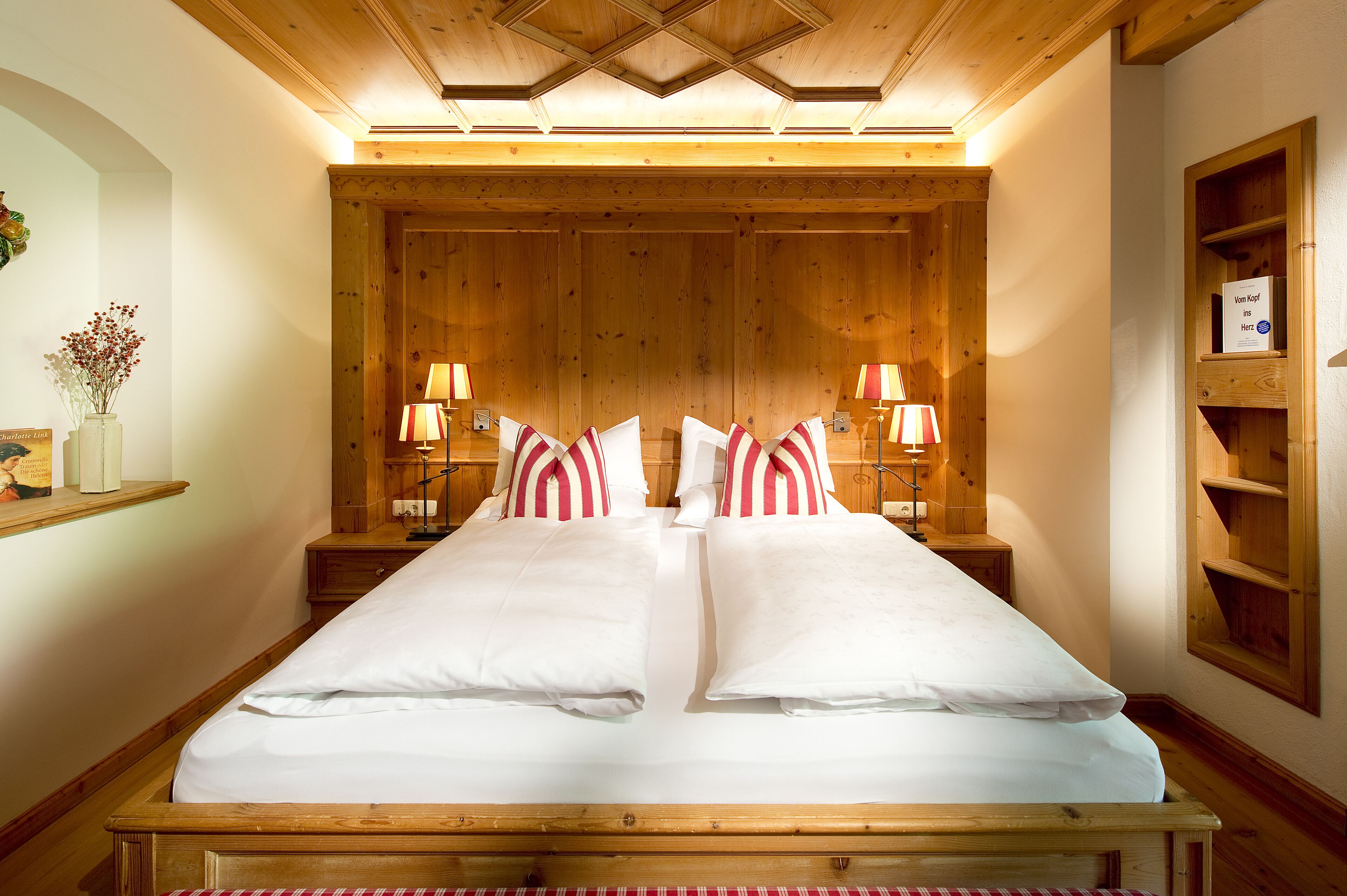 A double bed with white linens and red and white striped pillows, wooden headboard, bedside tables with lamps, and a wooden shelf with a sign.