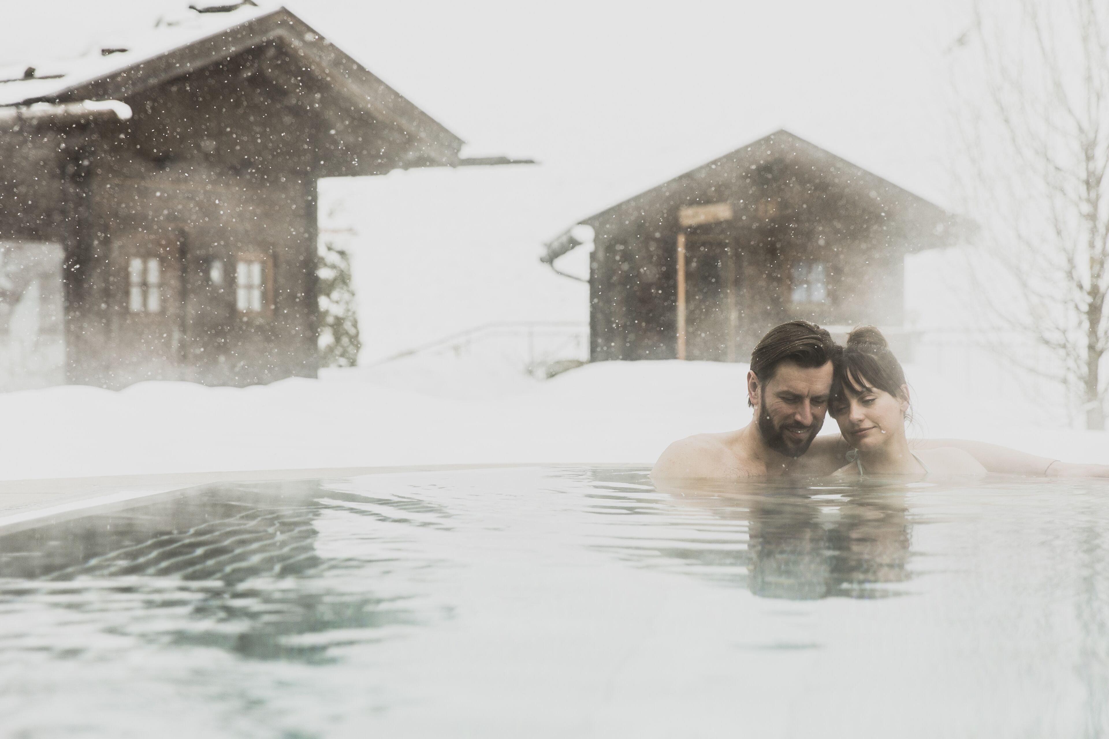 Una pareja en un jacuzzi con casas cubiertas de nieve al fondo.