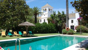 A swimming pool with lounge chairs and a thatched umbrella, surrounded by lush greenery and a white building with a bell tower. - Hotel Cortijo el Esparragal (Gerena)
