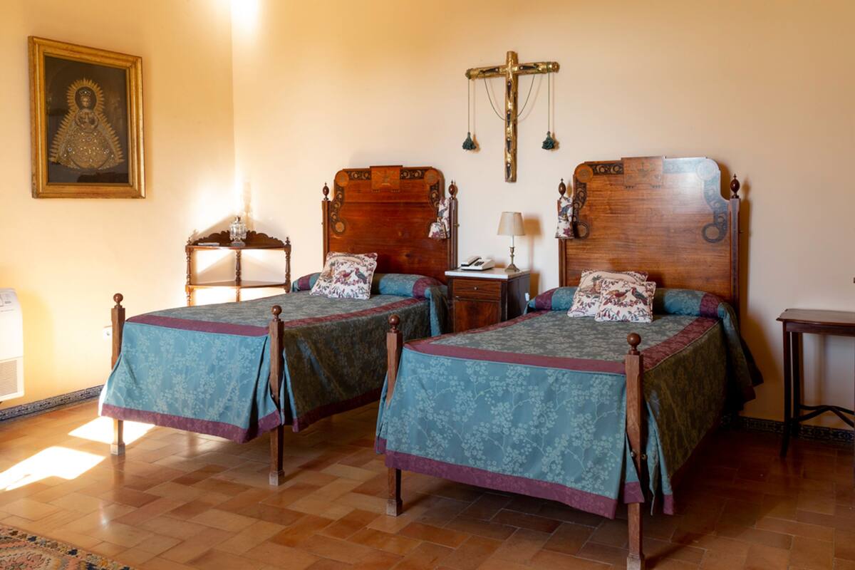 Une pièce comprenant deux lits en bois, un crucifix en bois, un tableau religieux encadré et une petite table avec une lampe.