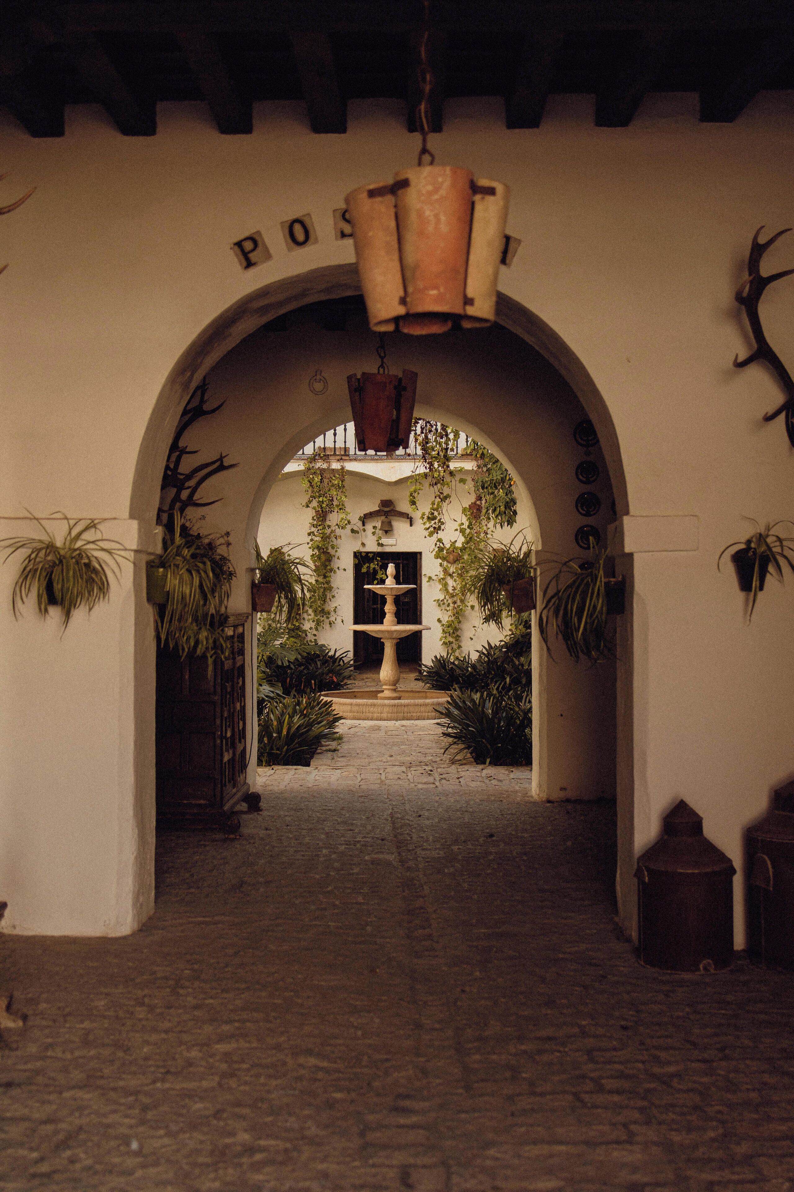 An arched entrance with a hanging light fixture, surrounded by potted plants and a stone pathway leading to a door.