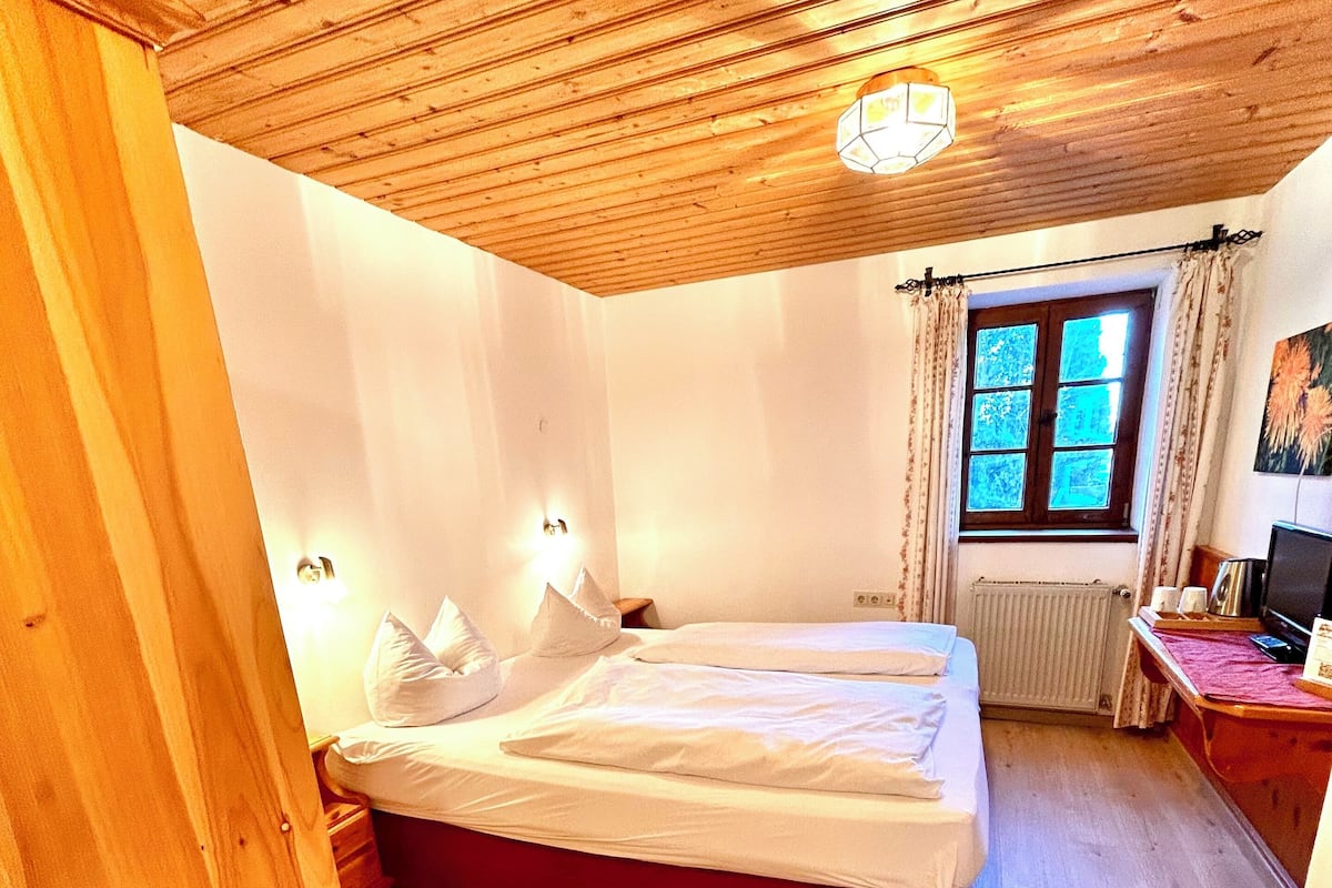 Een slaapkamer met een houten plafond, een bed met wit beddengoed, een radiator, een raam met gordijnen en een bureau met een computer.