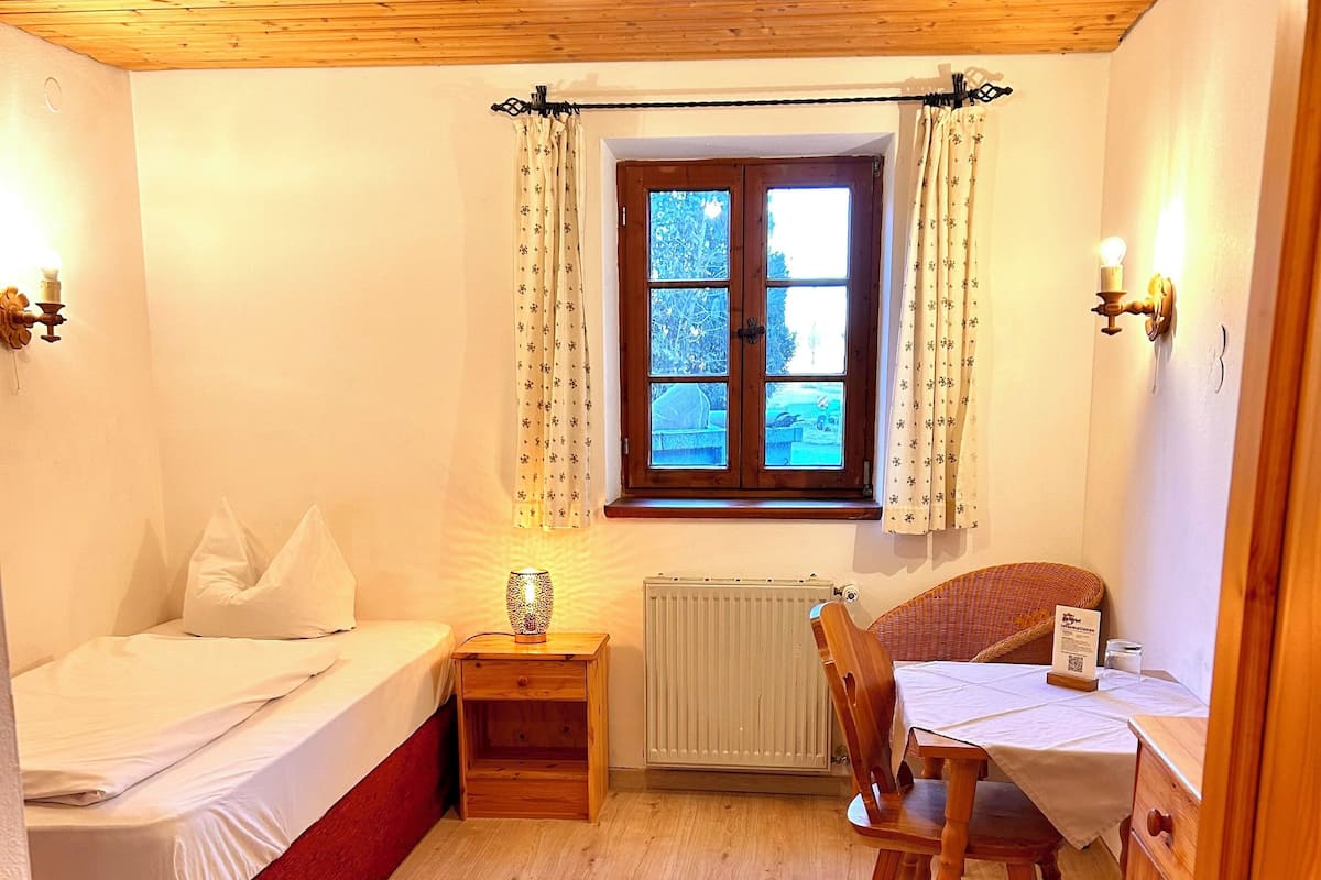Een knus kamertje met een houten plafond, een bed, een bureau en een stoel. Er is een raam met gordijnen en een radiator aan de muur.