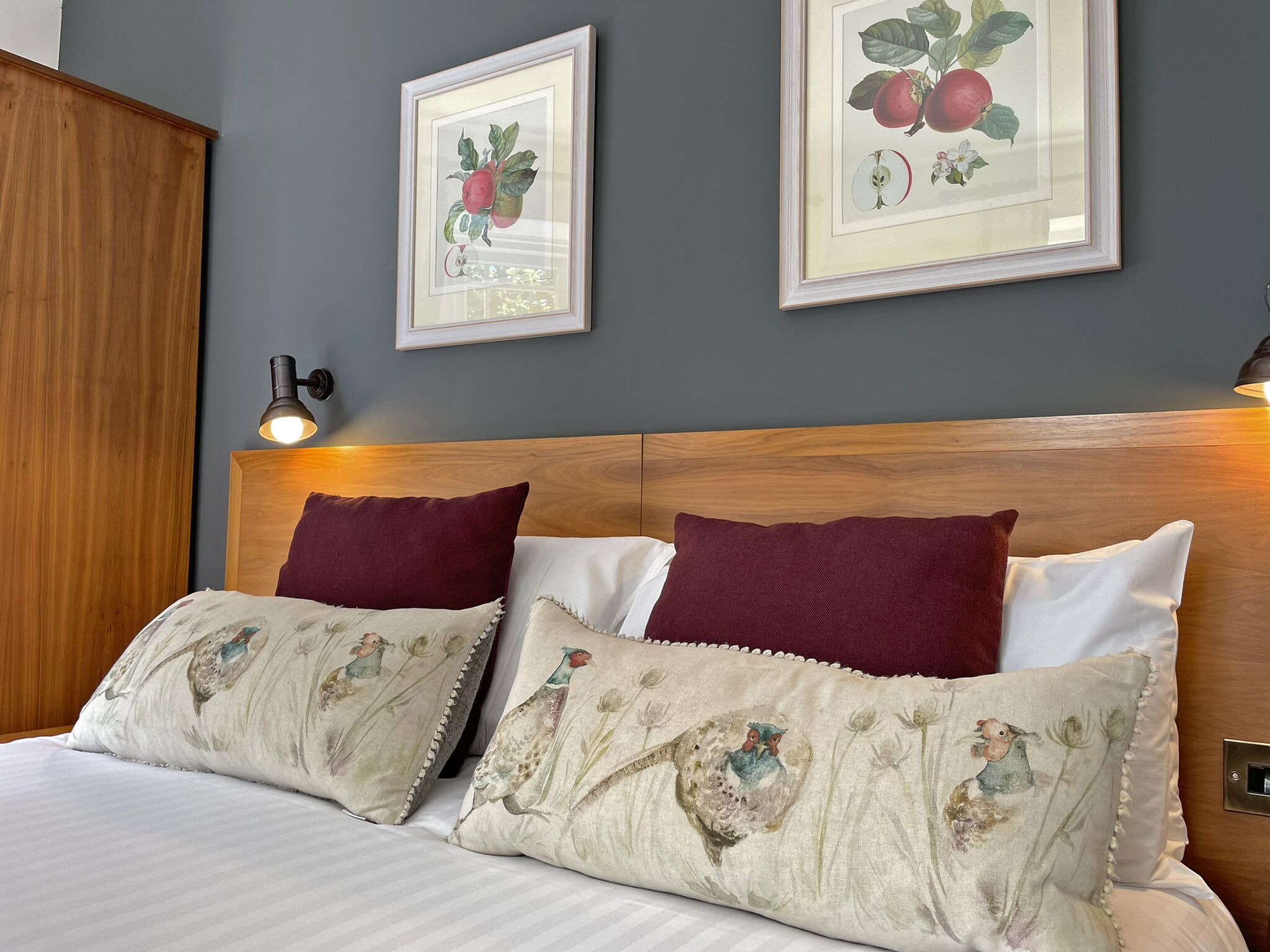 A neatly made bed with patterned pillows, a wooden headboard, and framed botanical prints on the wall.