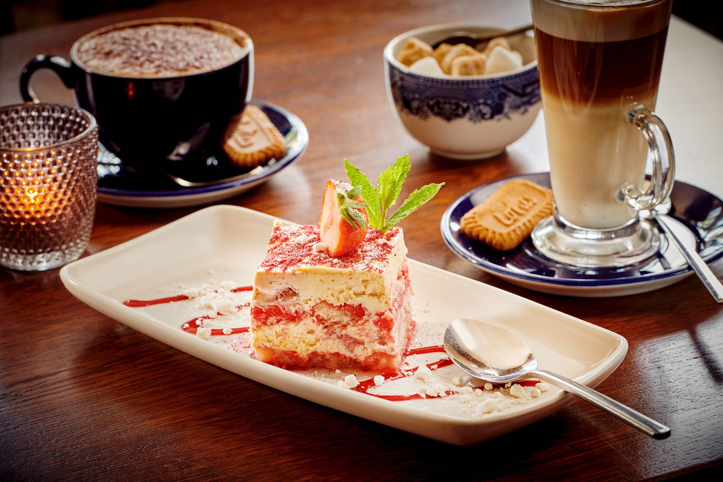 A dessert plate with a layered cake, a cup of coffee, and a bowl of cookies on a wooden table.