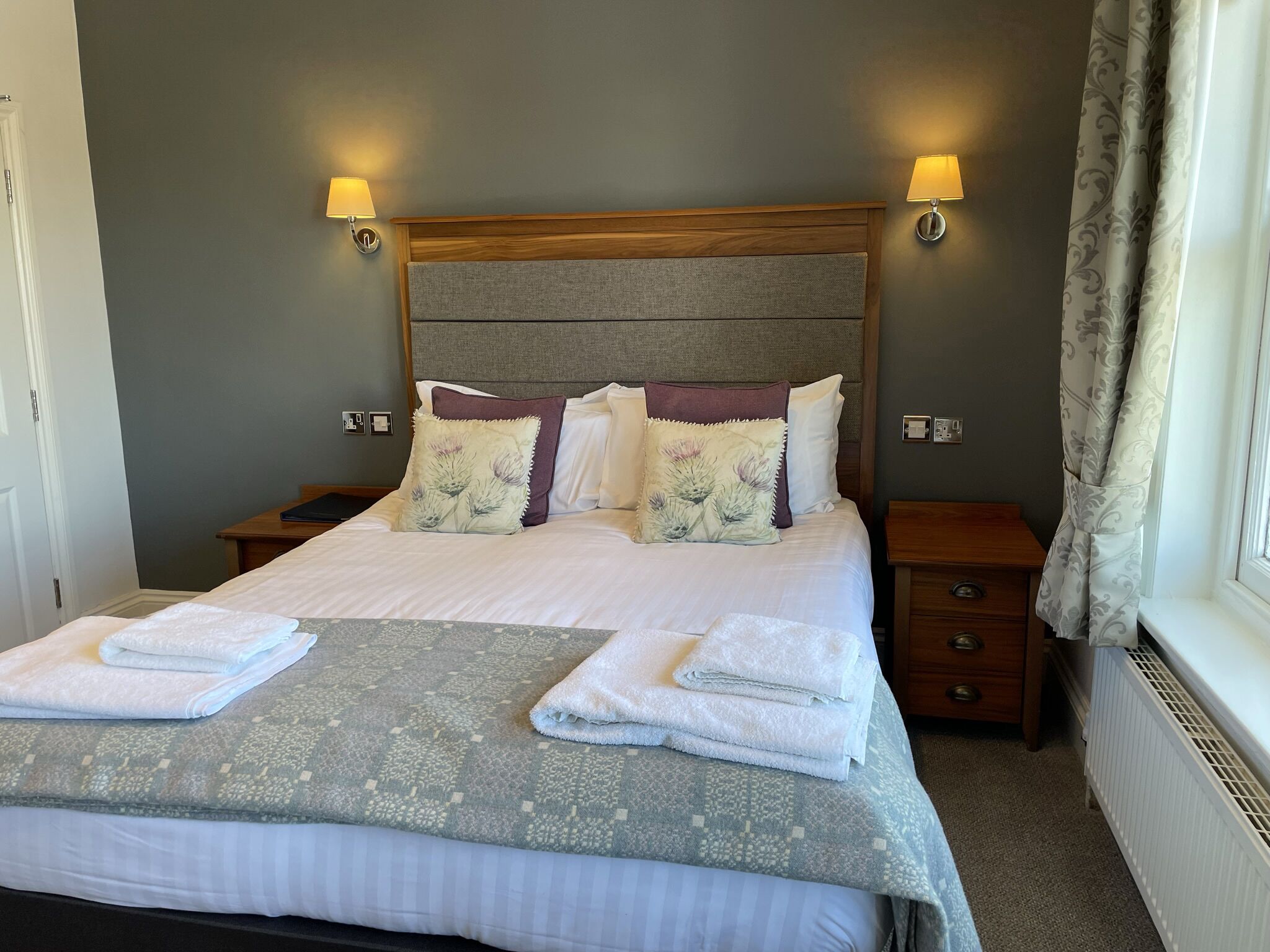 A neatly made bed with a grey and white patterned bedspread, two pillows with floral designs, and two folded white towels on the bed.