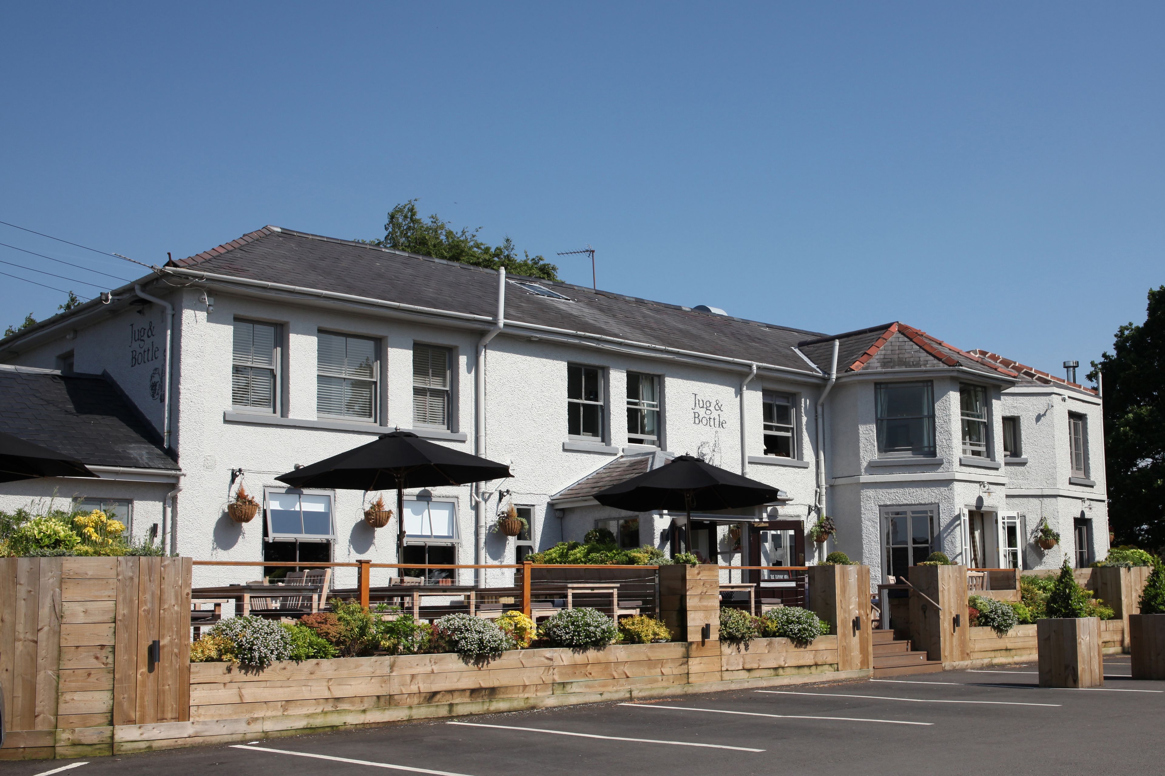 A two-story building with a sign reading "Jug & Bottle" on the facade. There are outdoor seating areas with umbrellas and a wooden fence surrounding the seating.