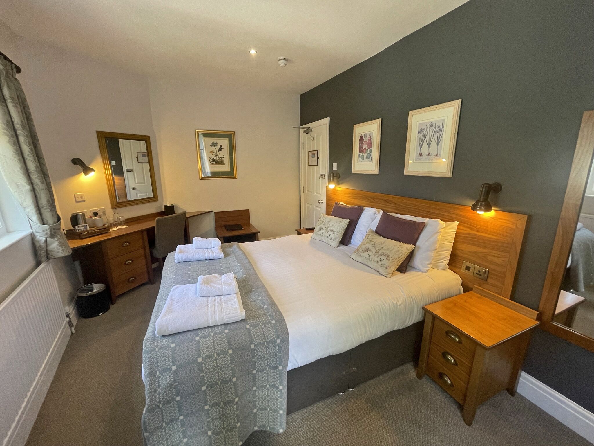 Habitación de hotel con una cama grande, mesitas de noche, un escritorio y una silla. Hay cuadros enmarcados en la pared y una ventana con cortinas.