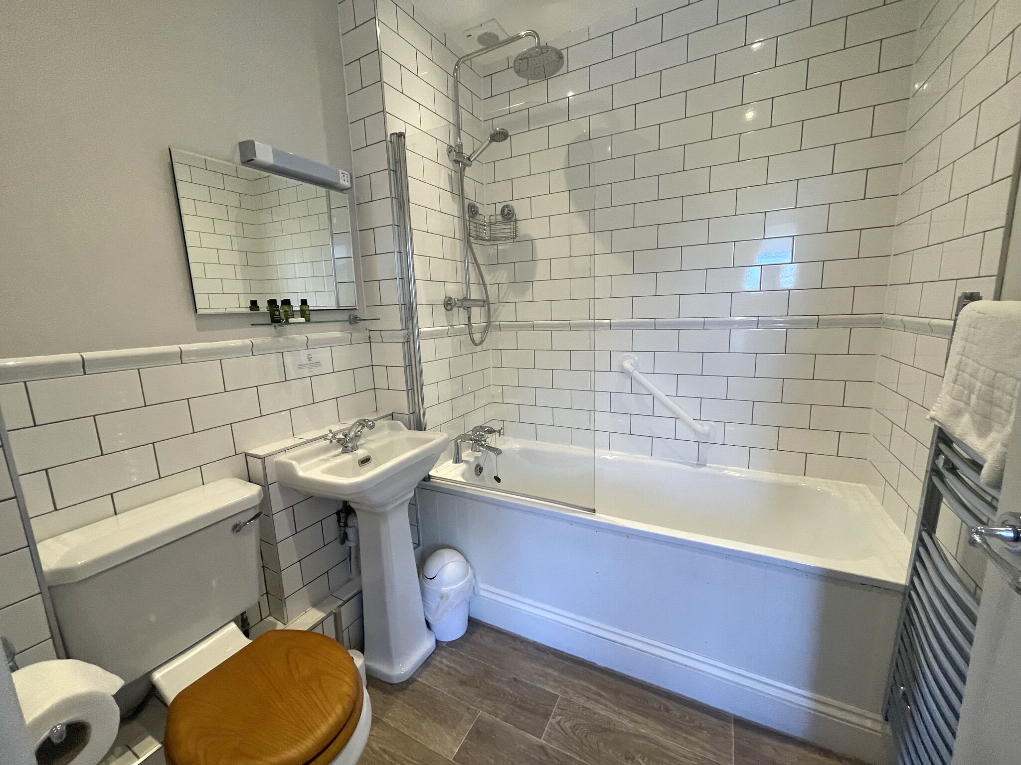 Un baño con azulejos blancos tipo metro, un lavamanos blanco, una bañera, un inodoro y un asiento de madera para el inodoro.