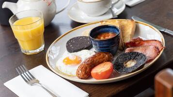 Un plato de desayuno con huevos, salchichas, tomates y morcilla, acompañado de una taza de café y un vaso de zumo de naranja.