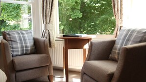 A cozy room with two armchairs and a small wooden table, a window with curtains, and a view of greenery outside. - The Jug & Bottle (Wirral)
