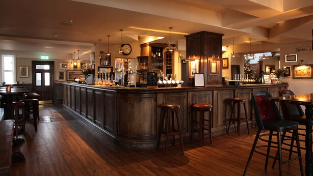 A bar area with wooden flooring, bar stools, and a counter with various bottles and glasses. - The Jug & Bottle (Wirral)