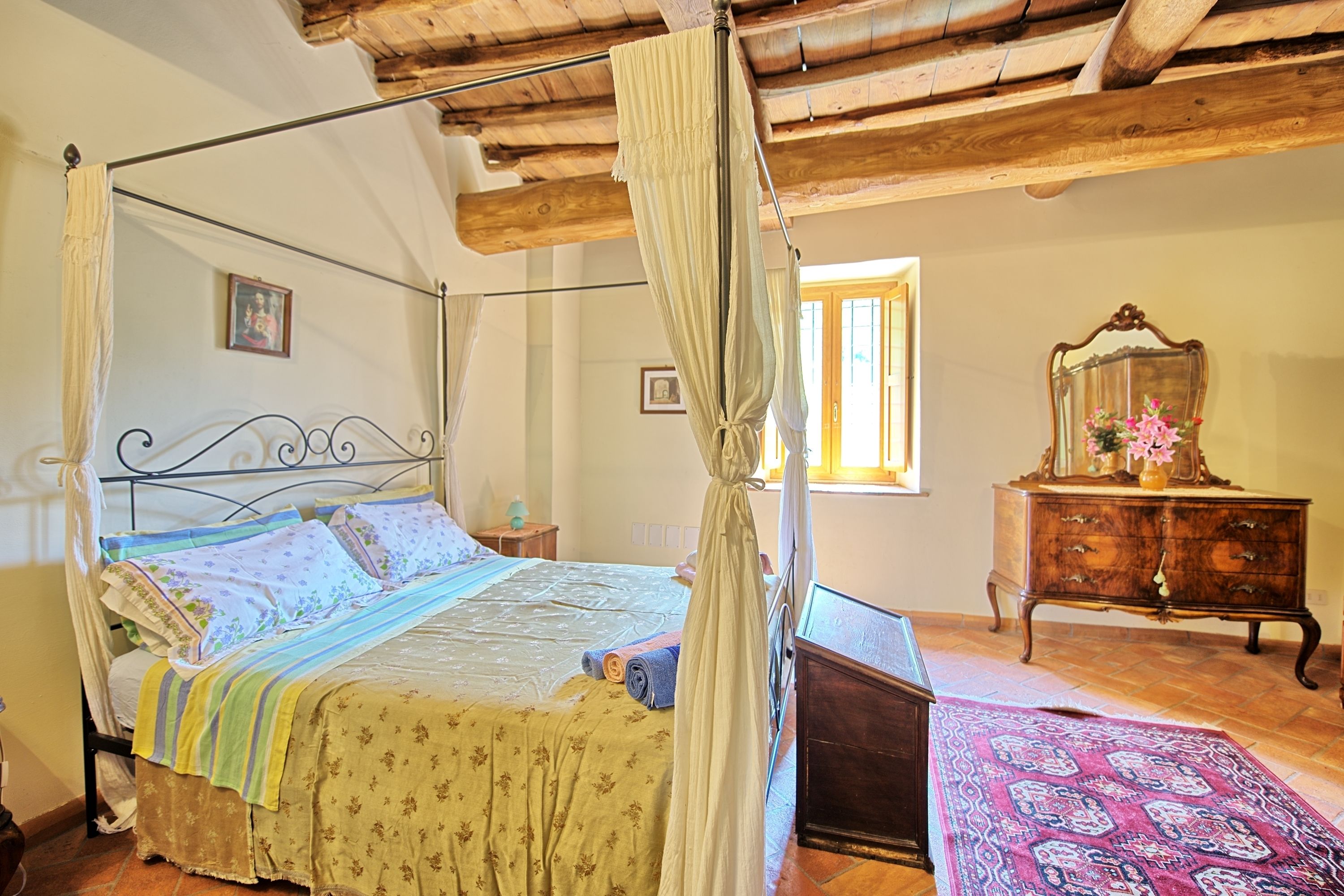 A four-poster bed with a canopy, a wooden dresser, and a patterned rug in a room with exposed wooden beams and a tiled floor.
