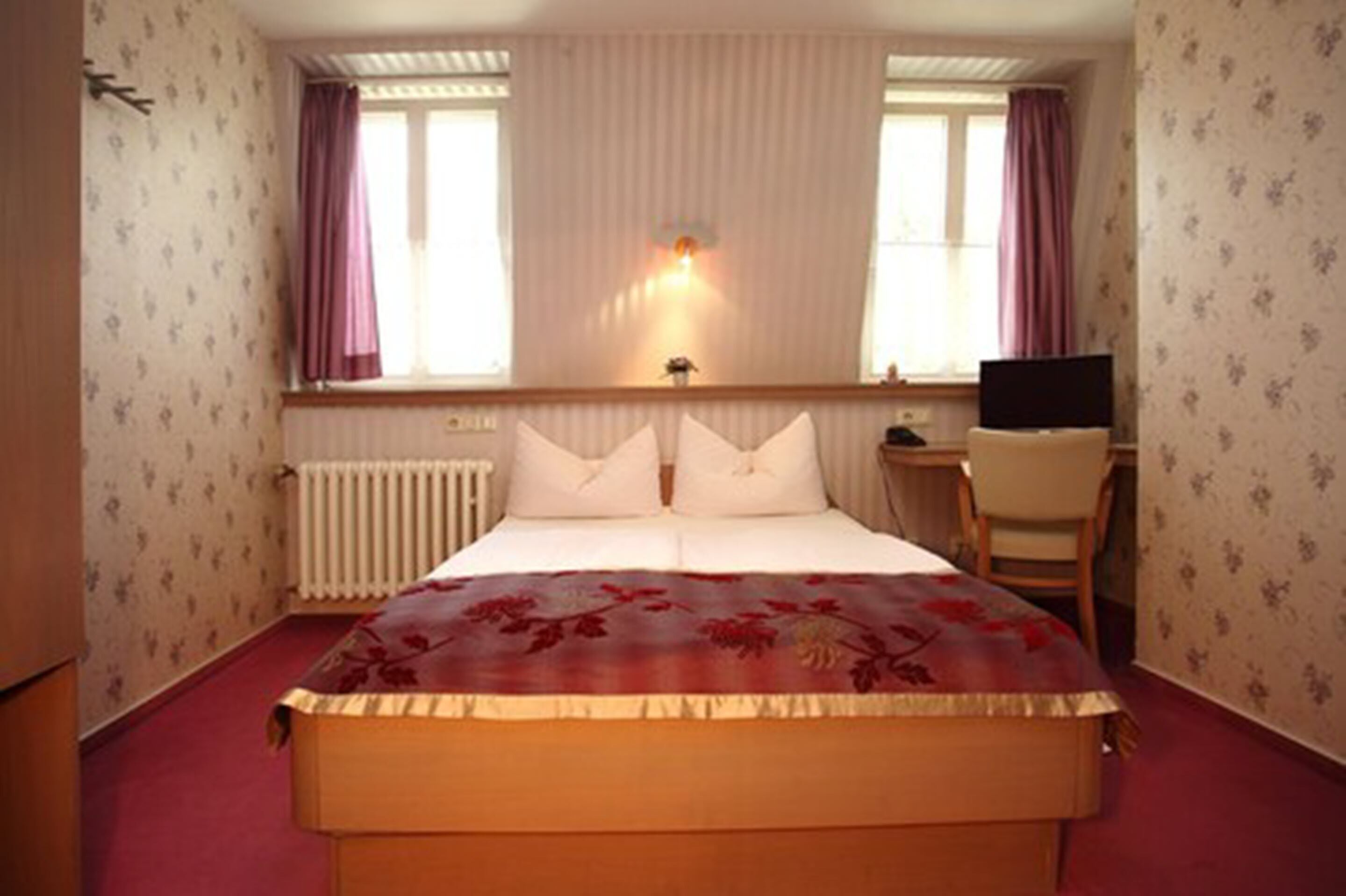 A hotel room with a bed, a desk, a chair, a radiator, and a window with curtains.