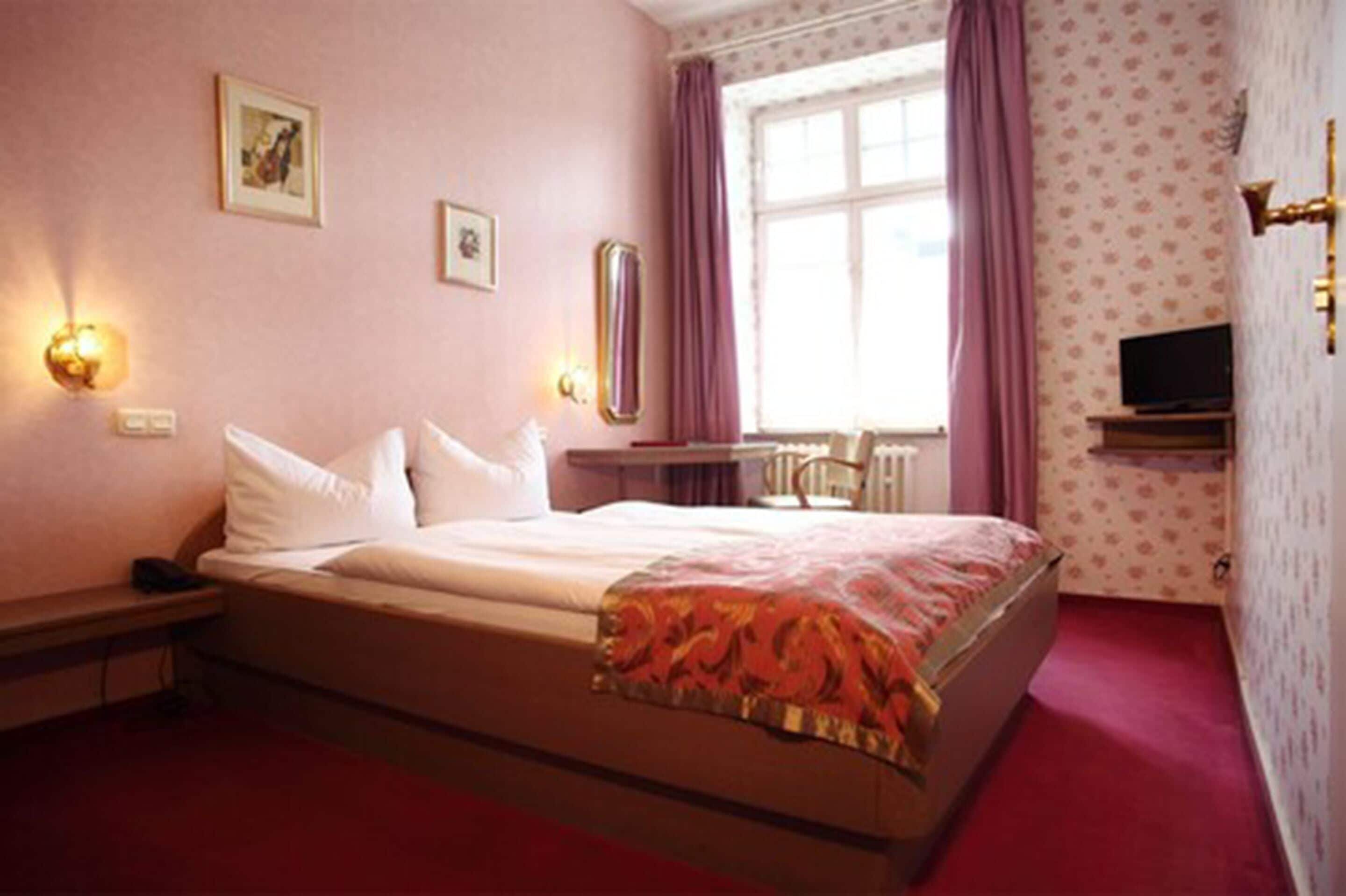 A hotel room with a bed, bedside lamps, a window with curtains, a television on a stand, and a radiator.