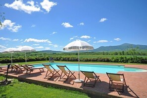 Ein Poolbereich mit Liegestühlen und Sonnenschirmen, umgeben von Grünflächen und Hügeln.