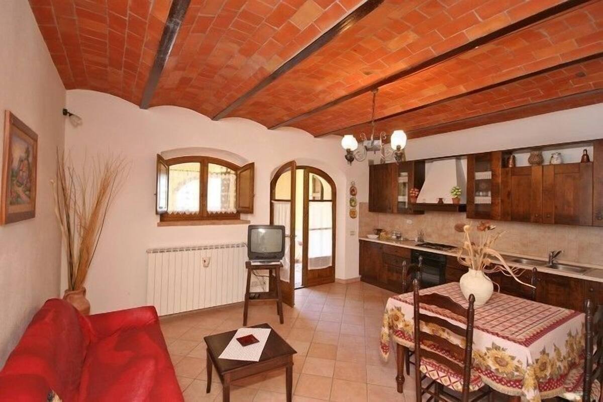 A cozy living room with a red sofa, a small table, and a TV. There's a kitchen area with wooden cabinets and a dining table set with a floral tablecloth.