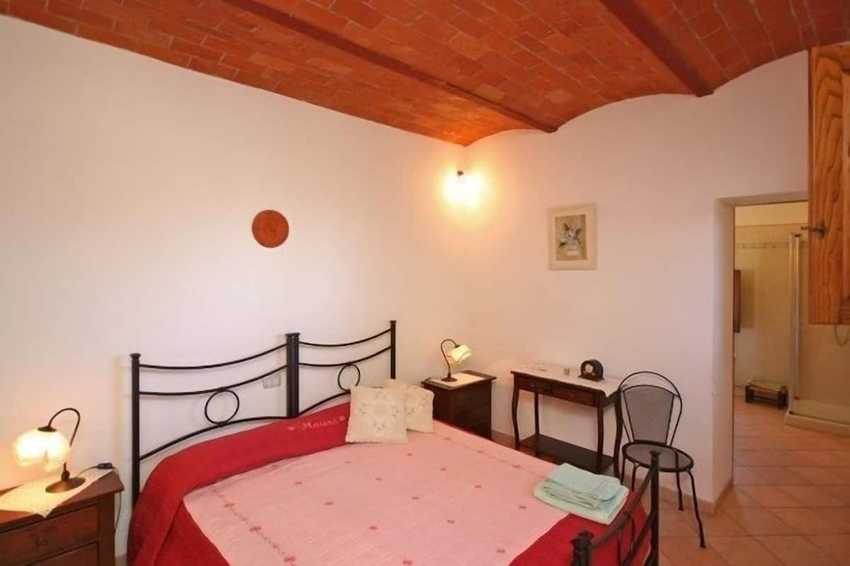 A bedroom with a bed, bedside table, lamp, desk, chair, and a small table with a lamp.