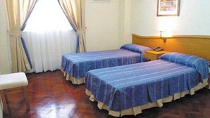 A room with two beds, a wooden bench, a nightstand with a lamp, and a window with curtains. - Alcor Hotel (Mendoza)