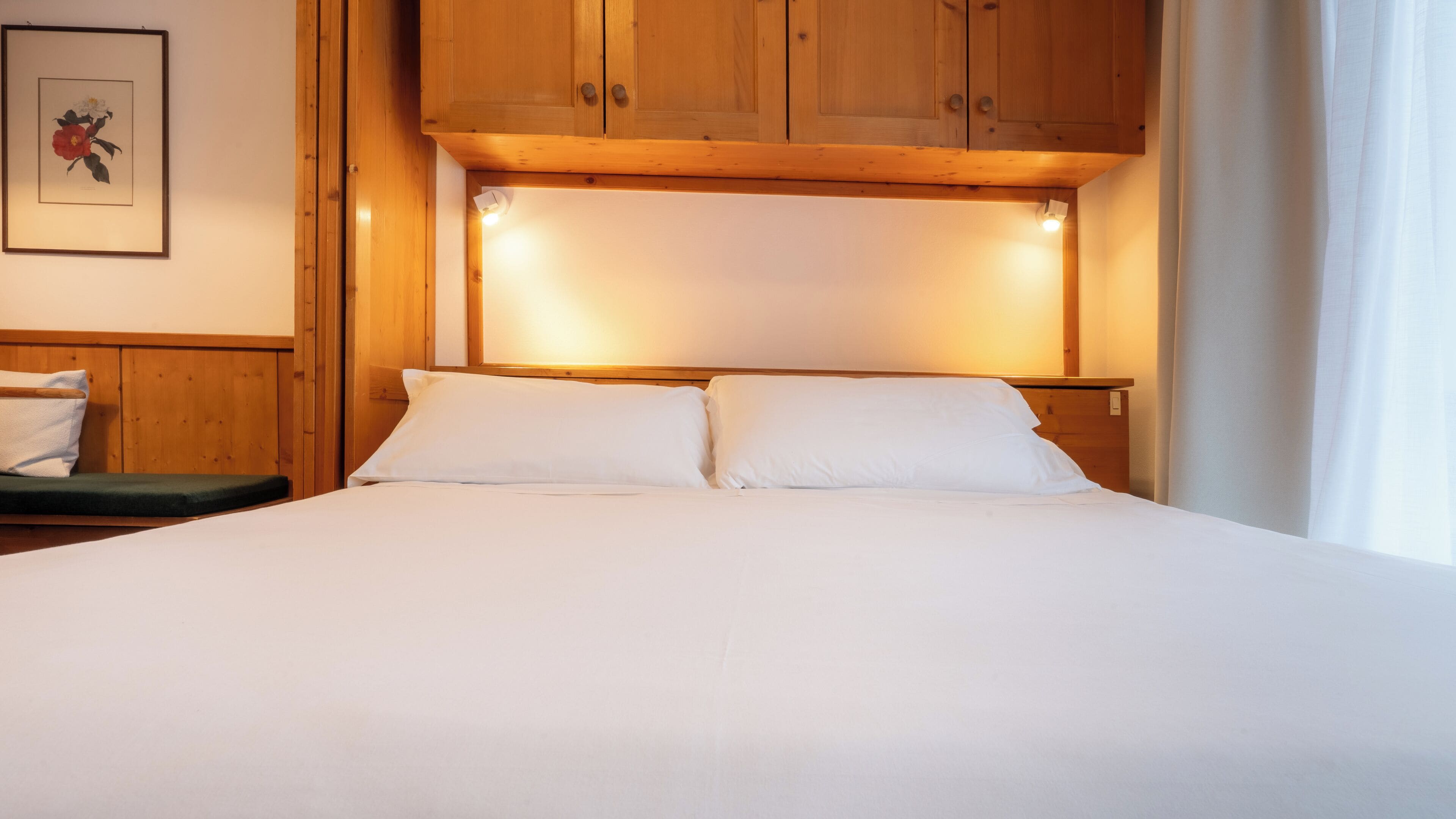 Un lit bien fait, avec du linge de lit blanc, une tête de lit en bois et une œuvre d’art florale encadrée accrochée au mur.