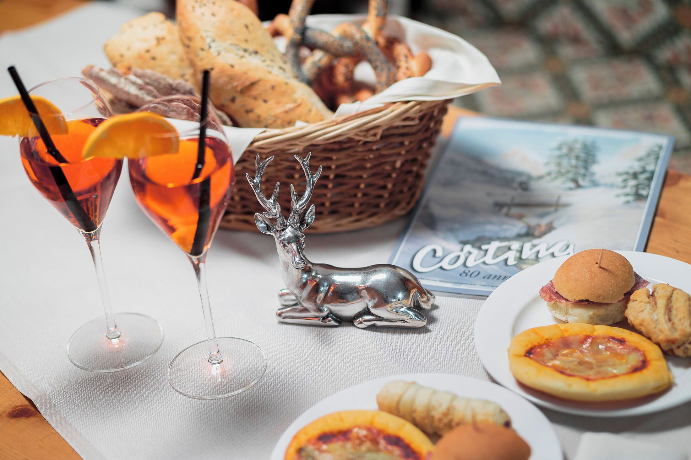 A table with a basket of bread, a plate with a sandwich, a plate with a pastry, and two glasses of a red beverage with orange slices.