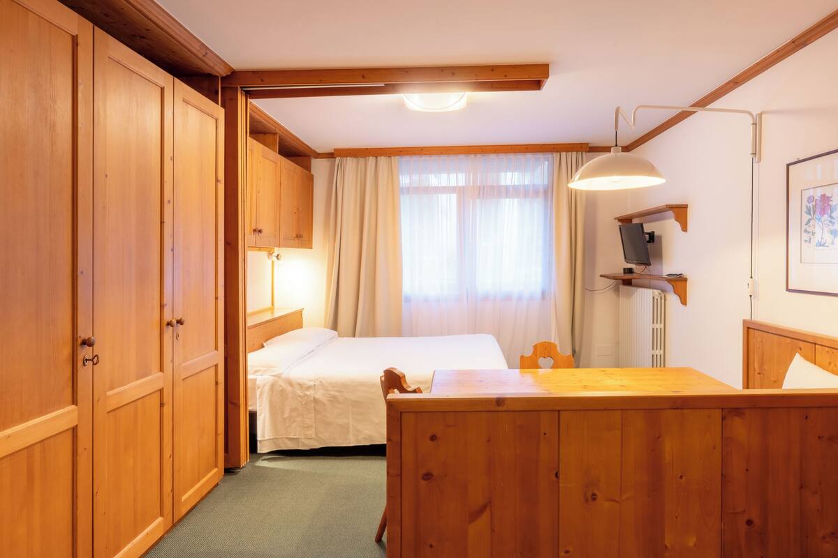 Une chambre d’hôtel avec des meubles en bois, un lit, un bureau, une chaise, une télévision et une fenêtre avec des rideaux.
