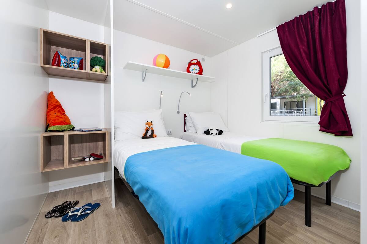 Een kamer met twee bedden, een raam met gordijn, een plank met diverse spullen, en een paar slippers op de vloer.