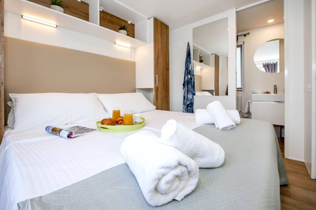 Een net opgemaakt bed met witte lakens, een groen dienblad met sinaasappelsap en croissants, en opgerolde witte handdoeken.
