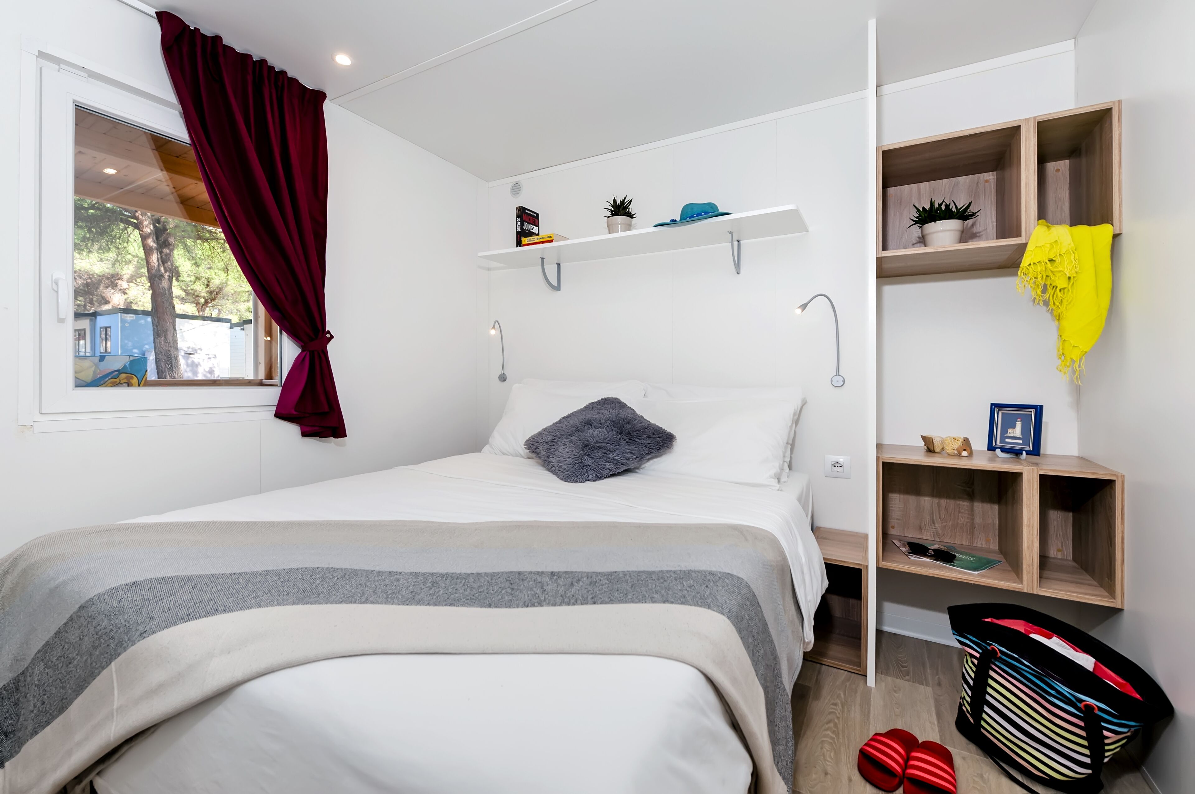 A neatly arranged bedroom with a bed, a window with curtains, a shelf with decorative items, and a small wooden cabinet.