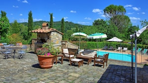 An outdoor patio with stone flooring, a stone fireplace, wooden furniture, and a pool area with umbrellas. - Casa Scopeti (San Casciano in Val di Pesa)