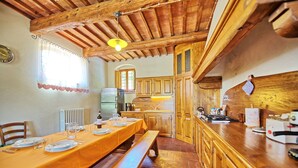 A rustic dining area with a wooden table set for a meal, a bench, and a kitchen with wooden cabinets and a brick floor. - Casa Scopeti (San Casciano in Val di Pesa)