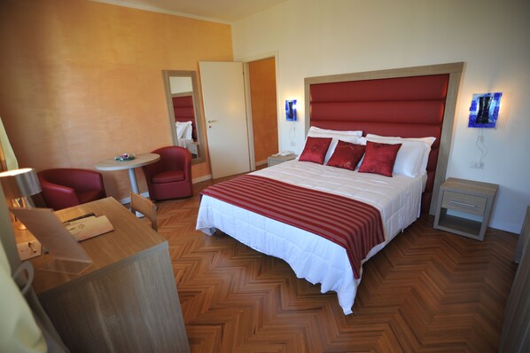 A hotel room with a large bed, a desk with a chair, a small table, and a mirror. - Hotel Astoria (Fermo)