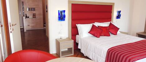 Quarto de hotel com cama, uma cadeira vermelha, mesa de cabeceira com telefone e luminária na parede.