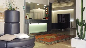 A reception area with a black leather sofa, a large potted plant, a reception desk, and a cactus in a white planter. - Hotel Astoria (Fermo)