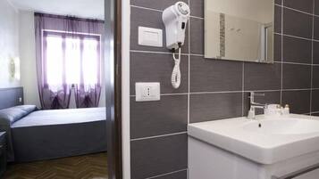 A hotel room with a bed, a bedside table, a window with curtains, a hairdryer on the wall, and a bathroom with a sink and mirror.