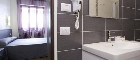 A hotel room with a bed, a bedside table, a window with curtains, a hairdryer on the wall, and a bathroom with a sink and mirror.